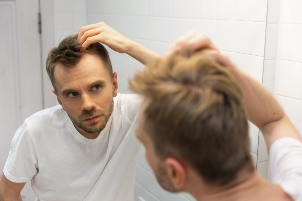 A man looks at his reflection in a mirror while examining his scalp and hair with his hand, for the best stylish haircut visit Orland Park Hair Salons.