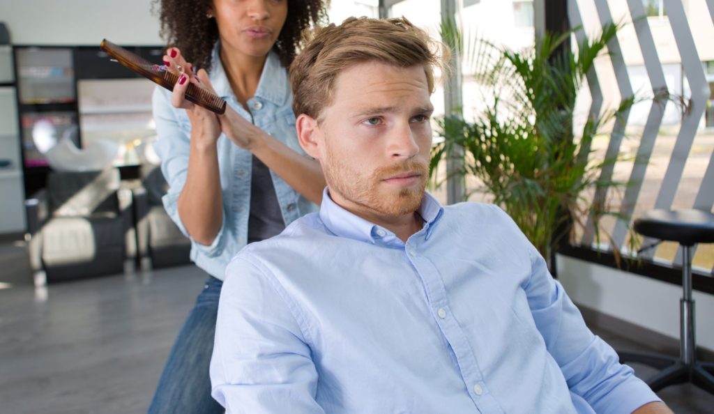How Do I Get the Exact Haircut I Want? A woman in a salon straightens a man's hair with a flat iron while he sits in a chair, looking ahead with a serious expression.