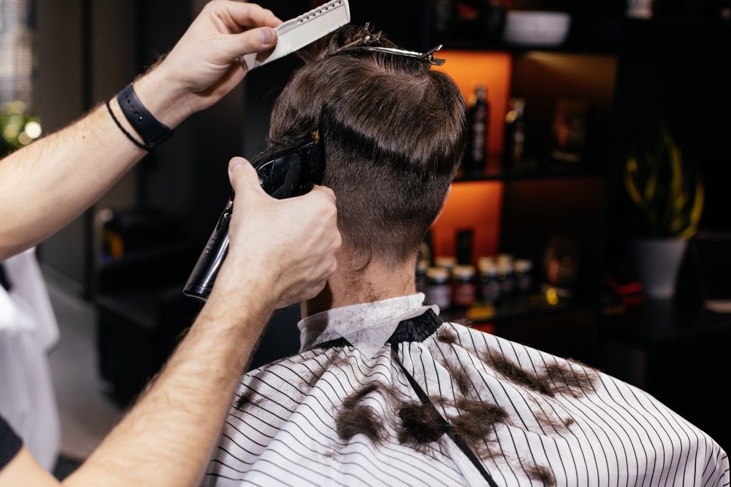 A person gets a haircut in an Oak Lawn barbershop, with a barber skillfully creating a hair fade using clippers and a comb to trim the back of their head. Hair clippings are visible on the striped cape.